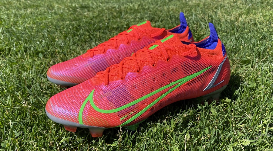 Red and neon green Nike Mercurial Vapor 14 boots placed on a natural grass field.