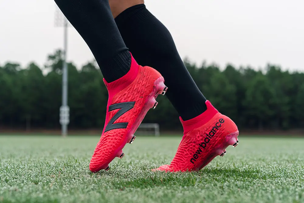 Bright red laceless New Balance Tekela v3+ boots being worn on a lush grass pitch.